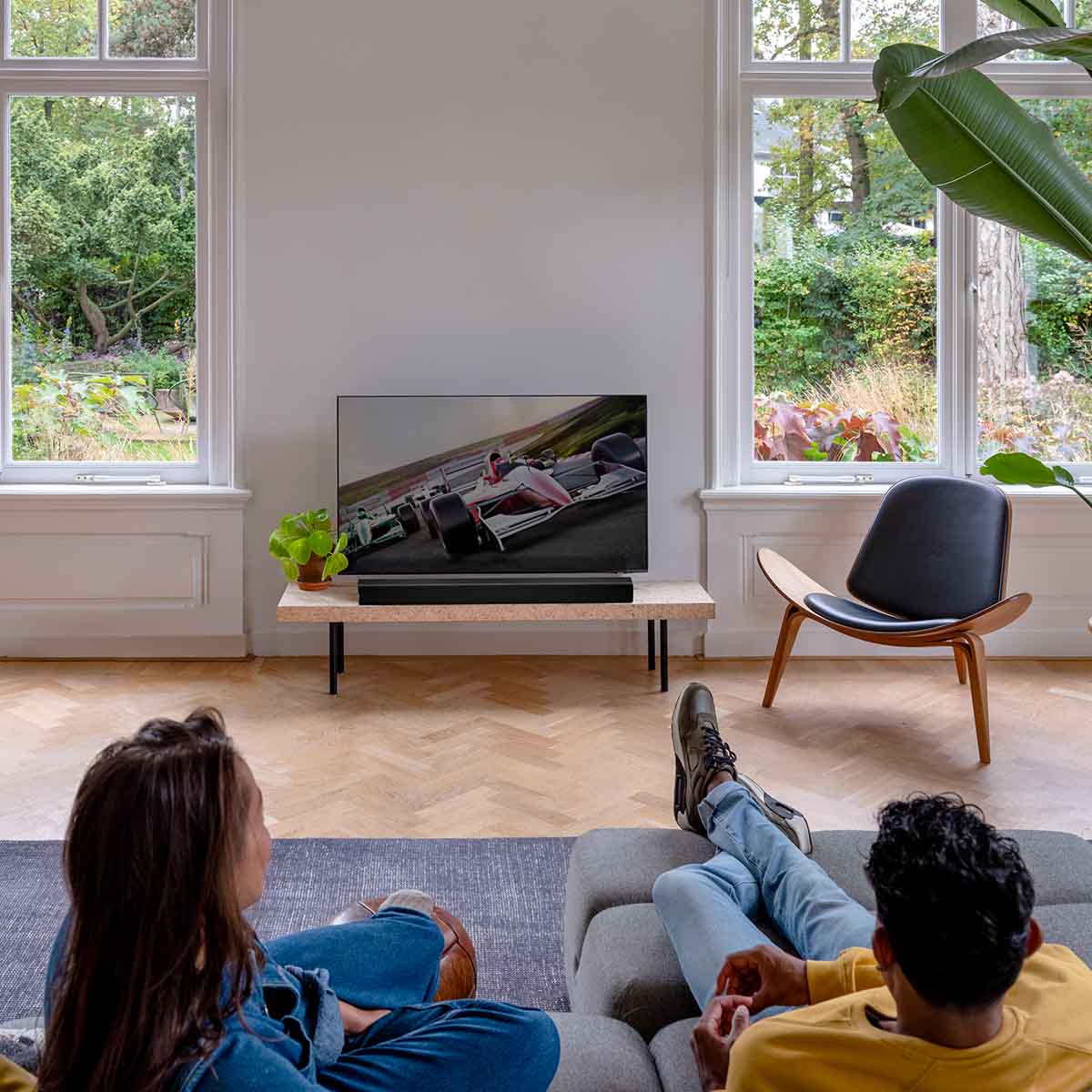 Harman Kardon Enchant 900 Soundbar lifestyle view with soundbar on table in front of TV and two people watching in foreground