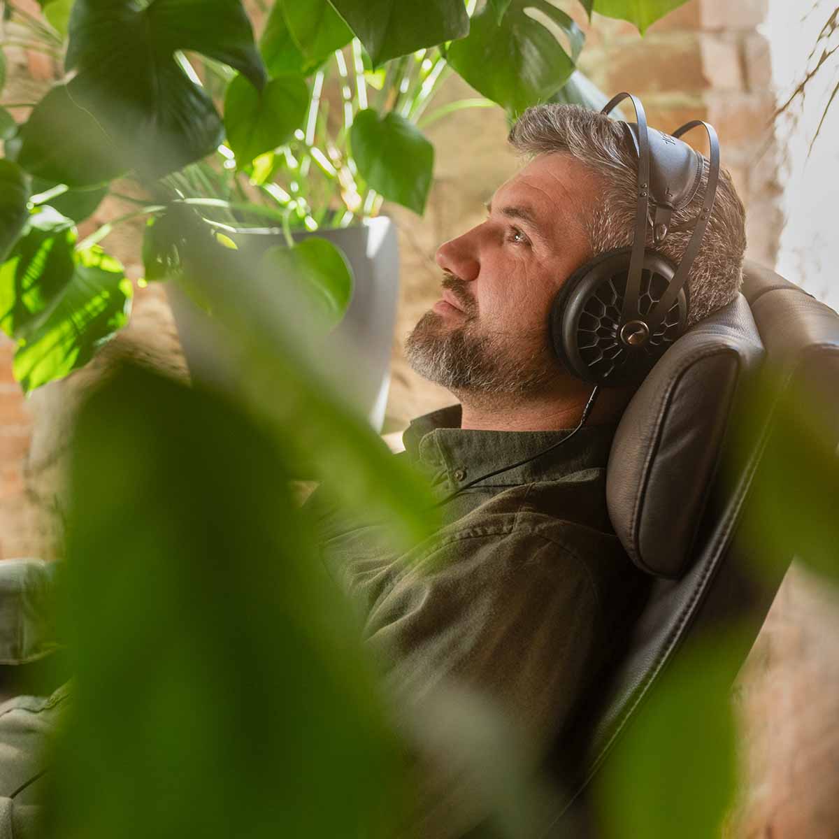 Meze 105 AER Open-Back Headphones - black lifestyle shot side view shot through plants of man wearing headphones reclining in a chair