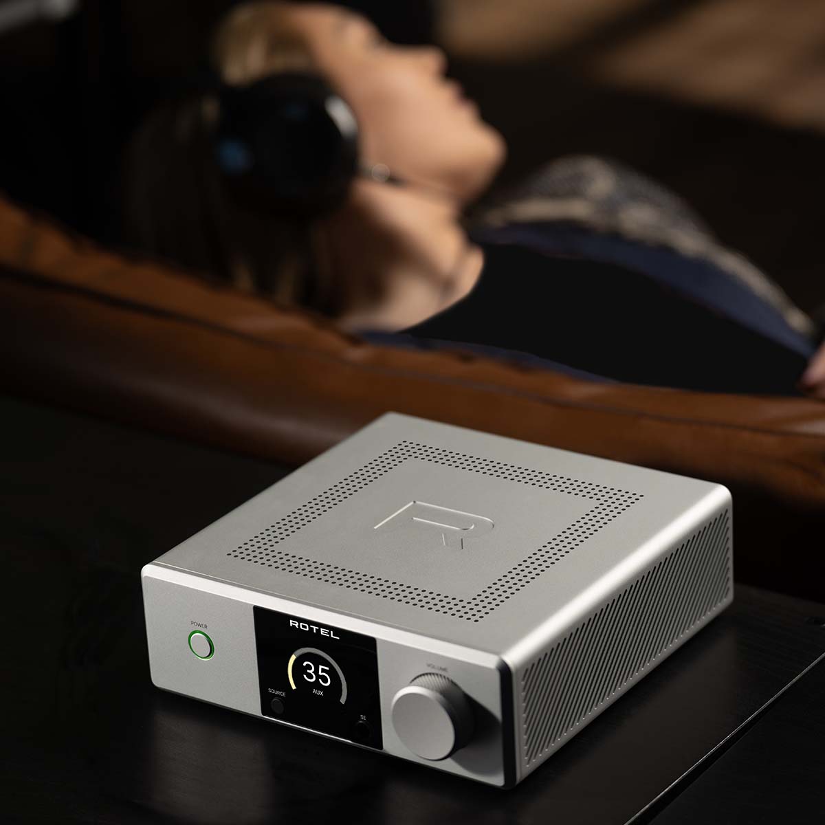 Lifestyle photo Rotel DX-5 Integrated Amplifier - silver top view angled left on cabinet with person listening to headphones out of focus in background