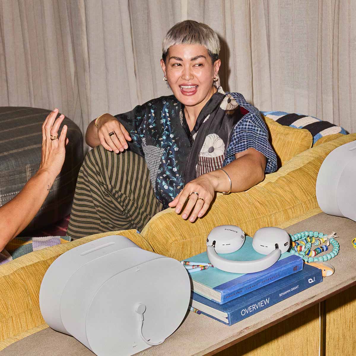Sonos Ace Ultra Wireless Over-Ear Headphones - lifestyle photo of a woman sitting on a couch with white headphones on a table behind the couch between other Sonos speakers