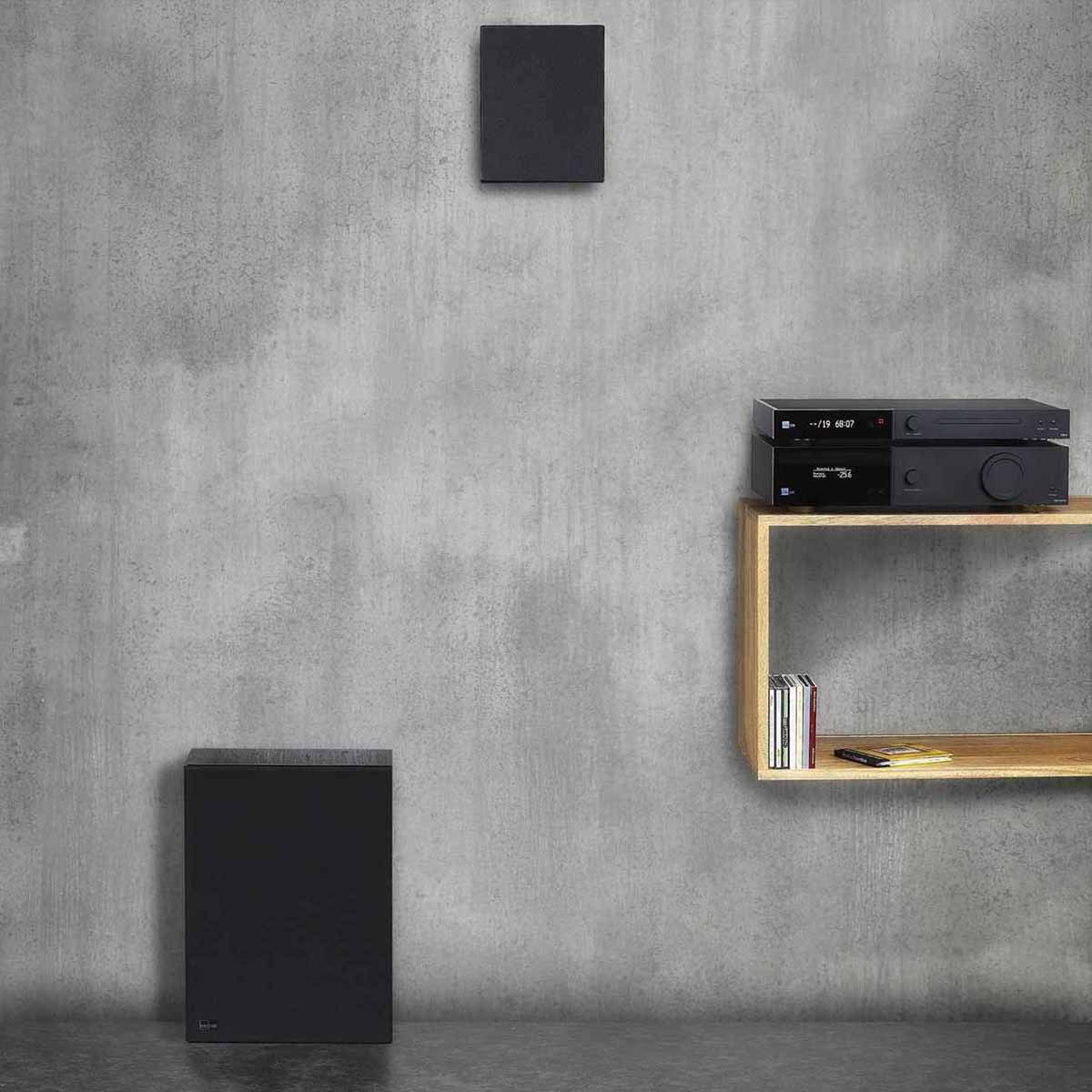 Lyngdorf BW-2 High Bandwidth Subwoofer black speaker with grille lifestyle view of speaker on floor with on-wall speaker above