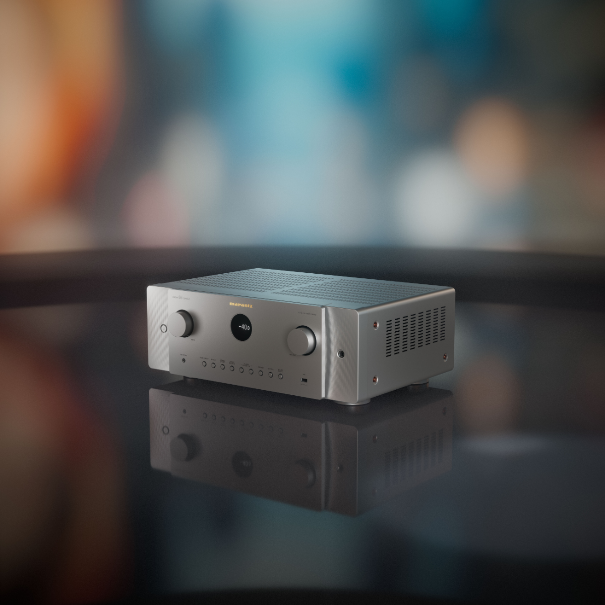 A silver metallic Marantz Cinema 60 Series 2 AV Receiver with two rotary knobs and a central circular dial, resting on a reflective dark surface against a blurred blue and orange background.
