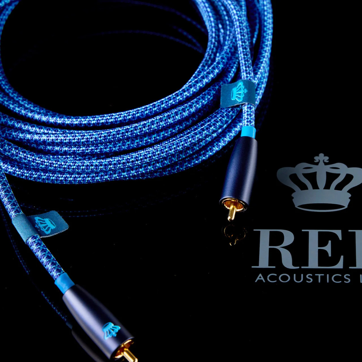A coiled blue braided REL Acoustics Commodore .1/LFE Subwoofer Cable with gold-plated connectors and small blue brand tags, photographed on a black reflective surface.