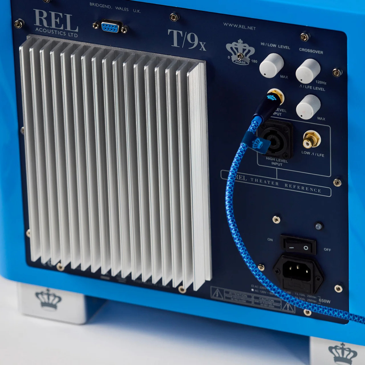 Close-up of the rear panel of a REL T9x audio amplifier, showing the silver heatsink, blue chassis, control knobs, and REL Acoustics Commodore .1/LFE Subwoofer Cable - Blue on a white background.
