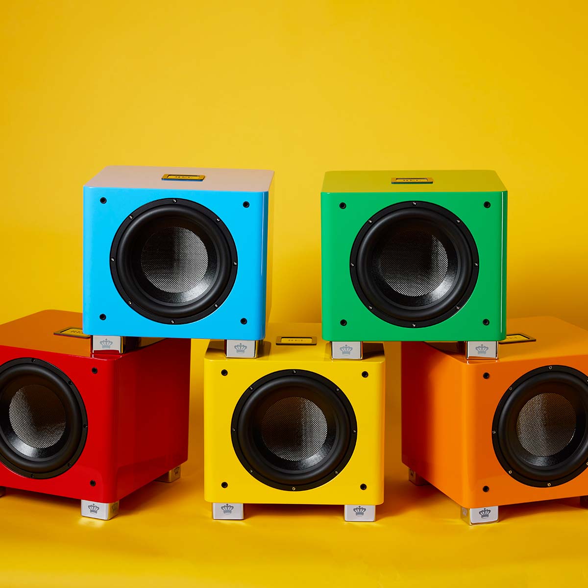 REL Acoustics T/9x SE Subwoofer pyramid of all five colors, blue and green stacked on red, yellow and orange all without grilles on yellow background