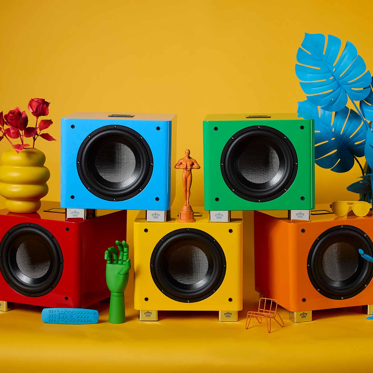 REL Acoustics T/9x SE Subwoofer pyramid of all five colors, blue and green stacked on red, yellow and orange all without grilles on yellow background with colored accessories surrounding units