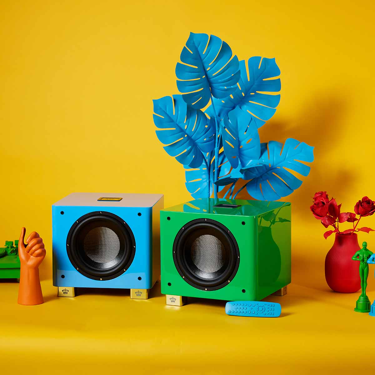 REL Acoustics T/9x SE Subwoofer Blue unit to left and Green unit to right front view without grilles on yellow background surrounded by colored accessories