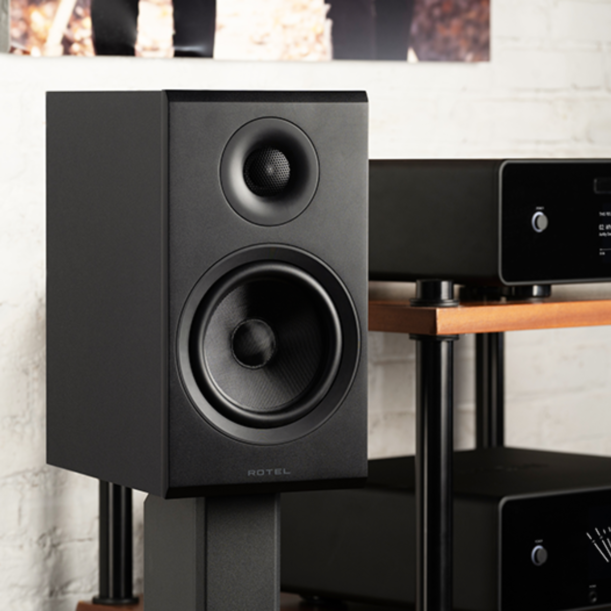 A black ROTEL Amora 5 bookshelf speaker shown in a home theater setting on a stand, next to an audio receiver on a wooden shelf.