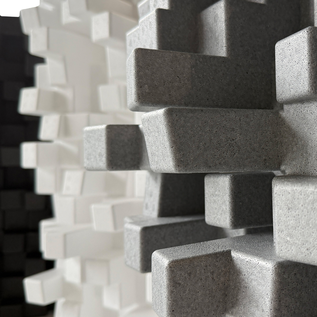 Close-up of a 3D geometric Vicoustic Cinema Multifuser DC4 Acoustic Diffuser Panel in a speckled gray finish, with white and black versions blurred in the background.