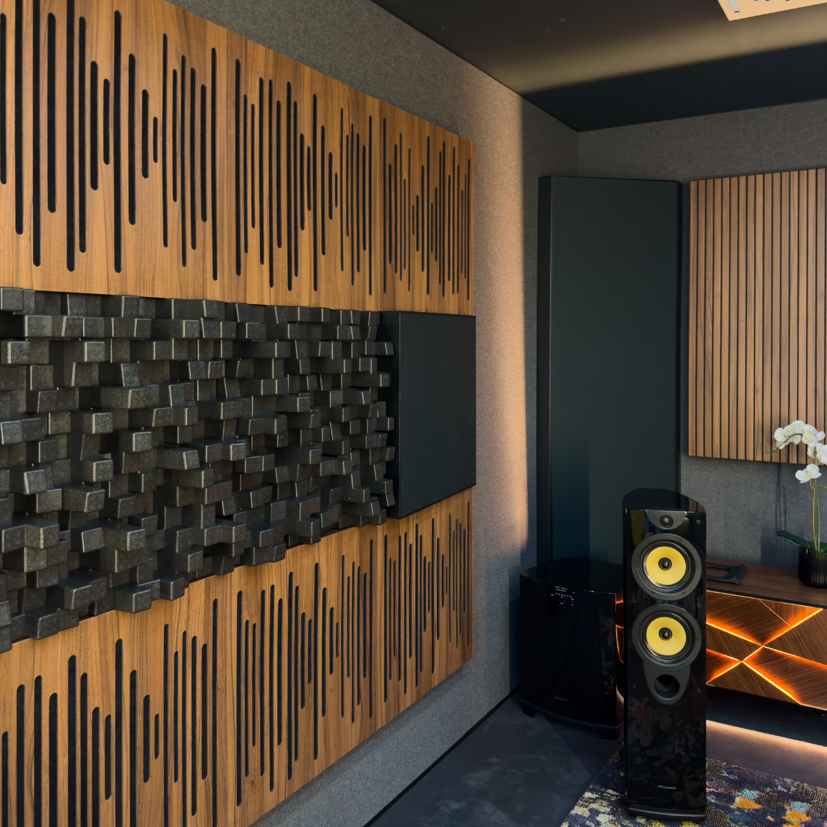 A modern room interior featuring a large wall-mounted speaker with two yellow cones, positioned in front of a dark gray cabinet. The background wall is decorated with Vicoustic Cinema Multifuser DC4 Acoustic Diffuser Panel.