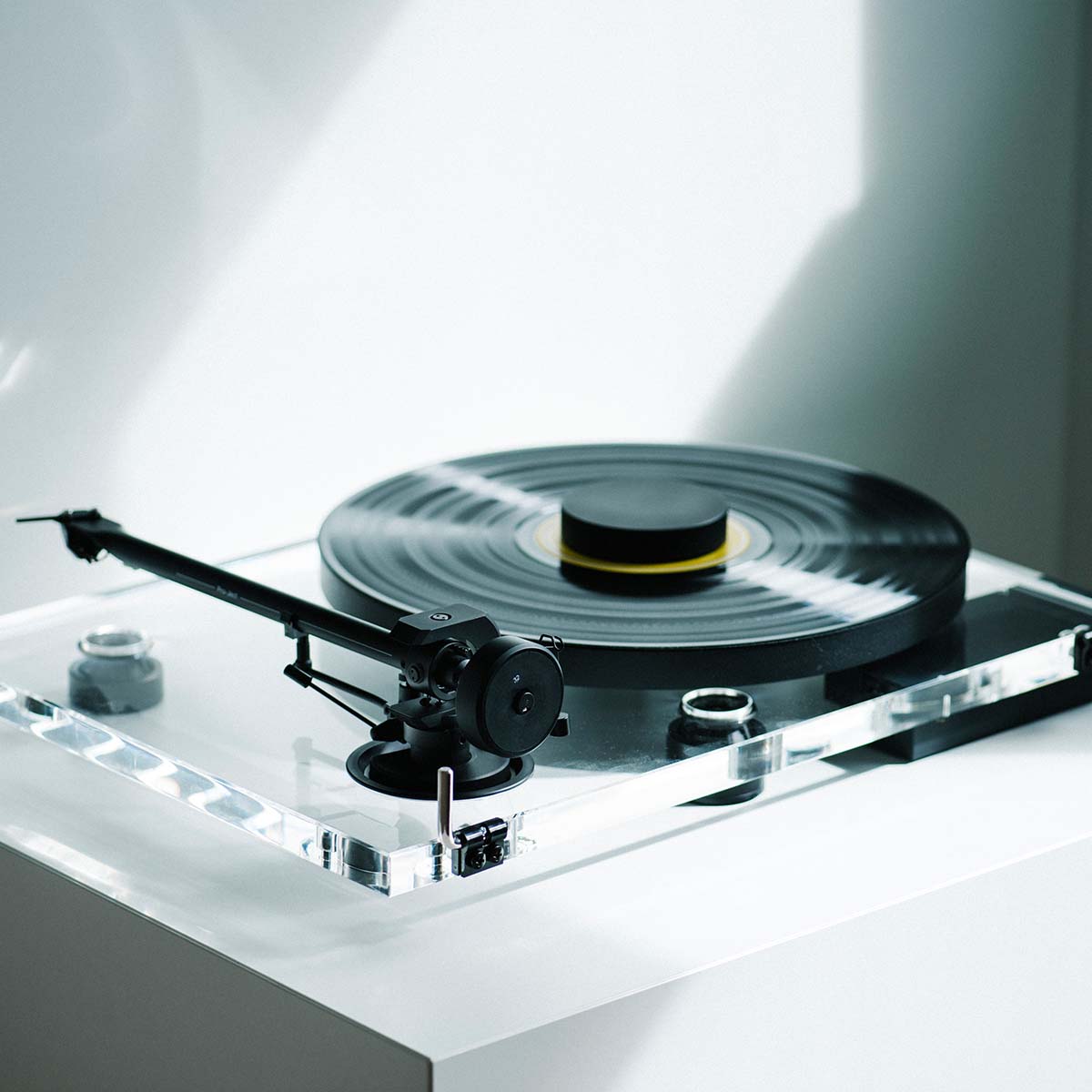 Pro-Ject XA B Balanced Acrylic Turntable - lifestyle shot high-angle rear view angled right of clear turntable on counter with sunlight reflected on back wall, record and record puck on the platter