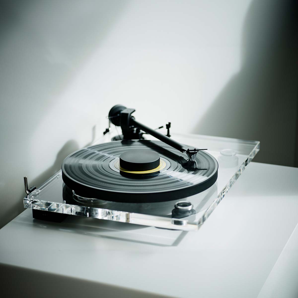 Pro-Ject XA B Balanced Acrylic Turntable - lifestyle shot high-angle side view angled right of clear turntable on counter with sunlight reflected on back wall, record and record puck on the platter and tone arm engaged