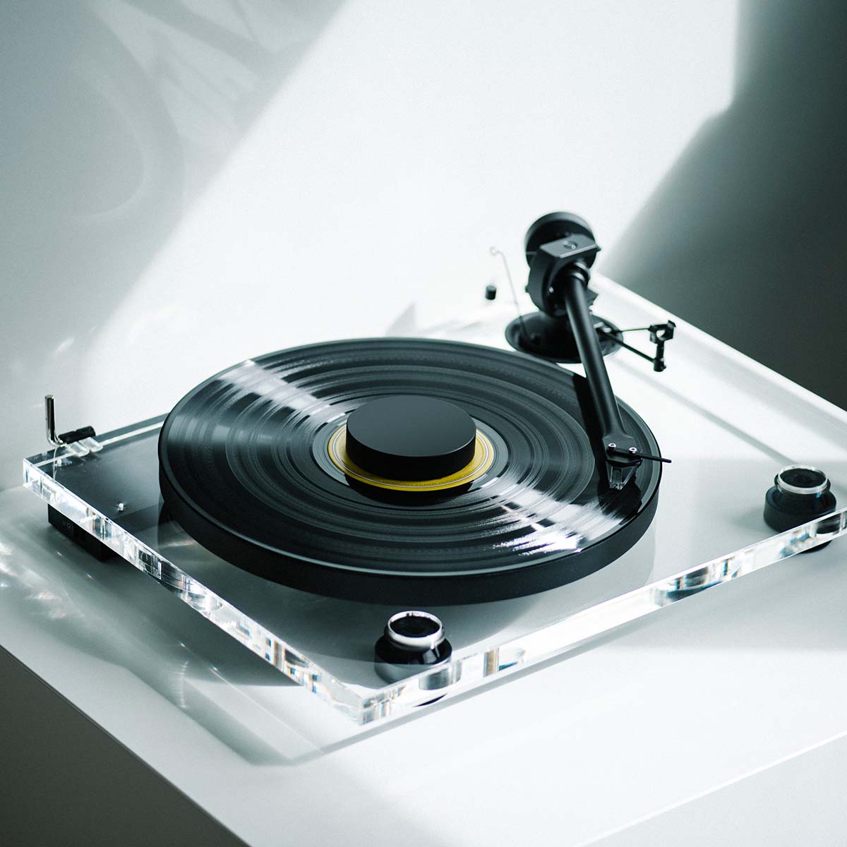 Pro-Ject XA B Balanced Acrylic Turntable - lifestyle shot high-angle front view angled right of clear turntable on counter with sunlight reflected on back wall, record and record puck on the platter and tone arm engaged