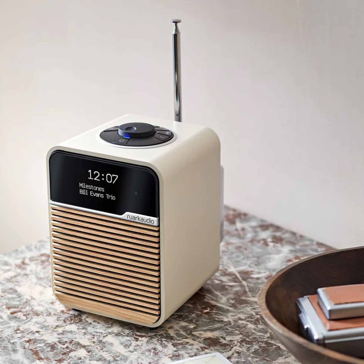 Ruark R1 Mk4 Deluxe Bluetooth Radio - Light Cream with Ash grille on marble counter with antenna raised - three-quarter front view angled left with time and song info on screen