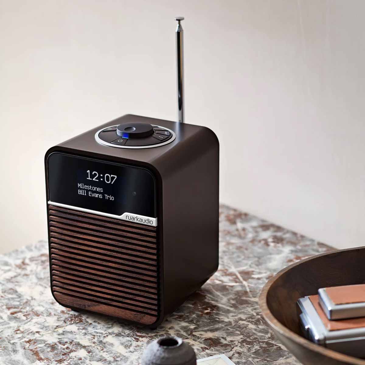 Ruark R1 Mk4 Deluxe Bluetooth Radio - Espresso with Walnut grille on marble counter with antenna raised - three-quarter front view angled left with time and song info on screen