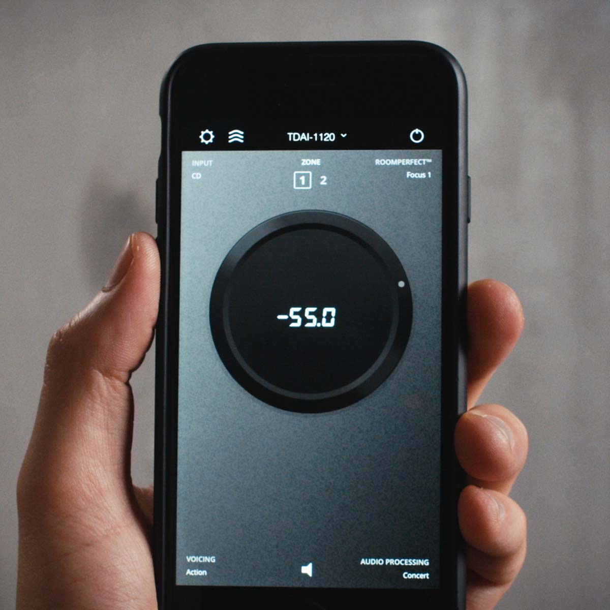 Hand holding a smartphone displaying an Lyngdorf RoomPerfect app on a gray background