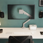Modern office desk setup with a monitor on a stand, keyboard, and speakers against a teal wall.