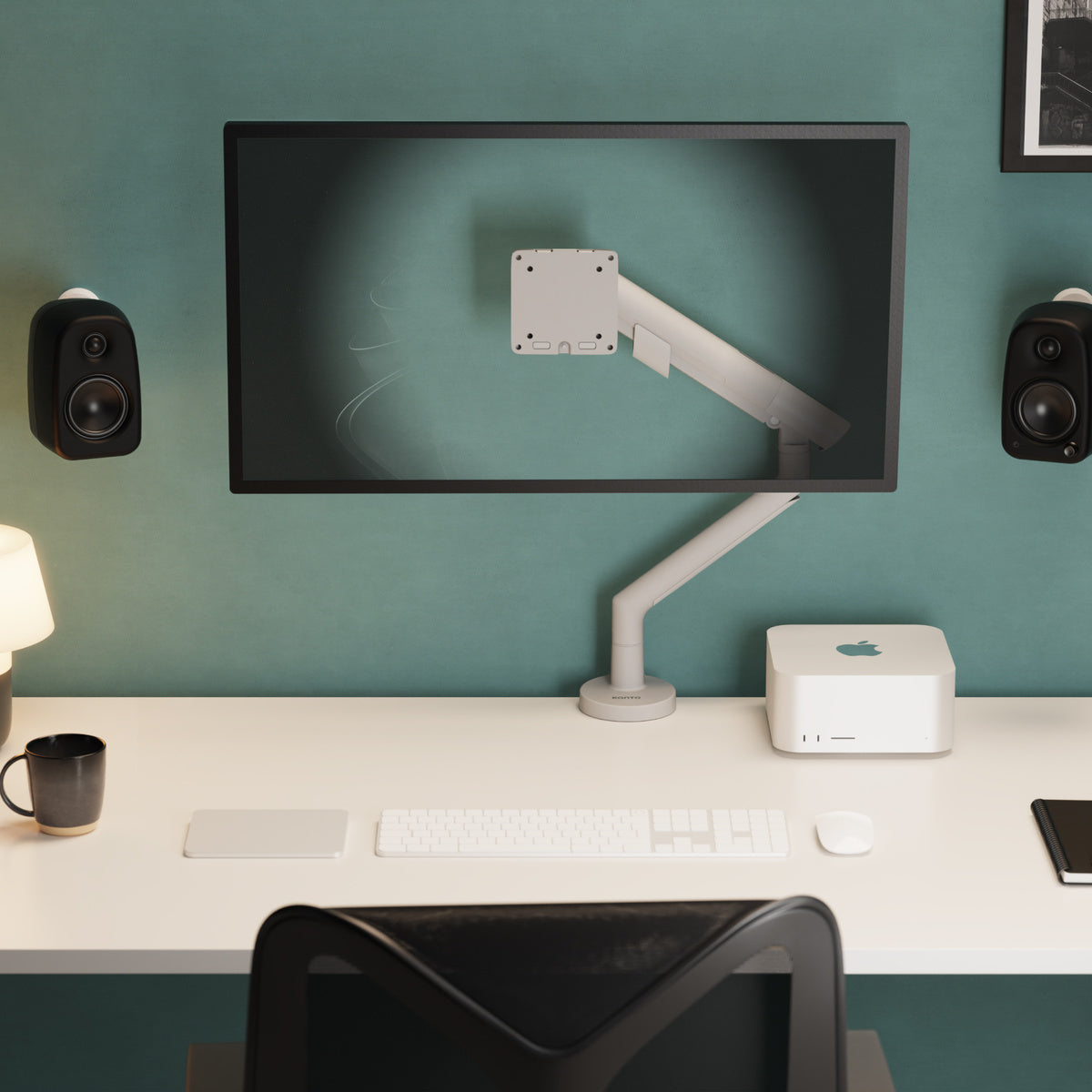 Modern office desk setup with computer monitor, keyboard, and speakers against a teal wall.