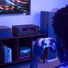 Denon AVR-S980H AV Receiver lifestyle photo of unit on cabinet under TV with person playing video games to front