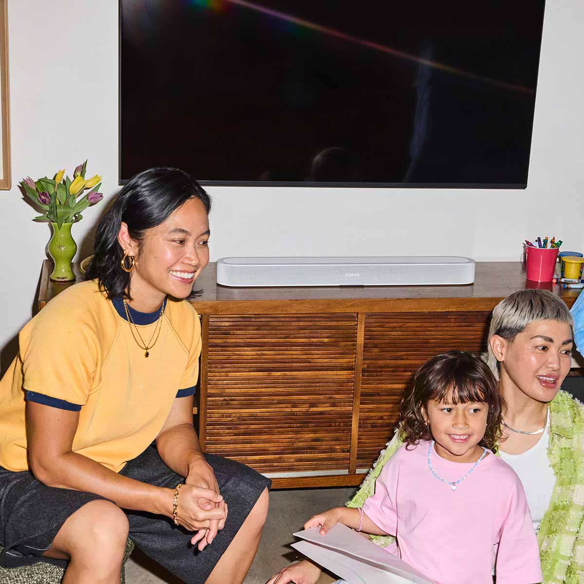 Sonos Beam Ultra Soundbar - lifestyle photo of white soundbar on a cabinet below a TV with two women and a girl in foreground