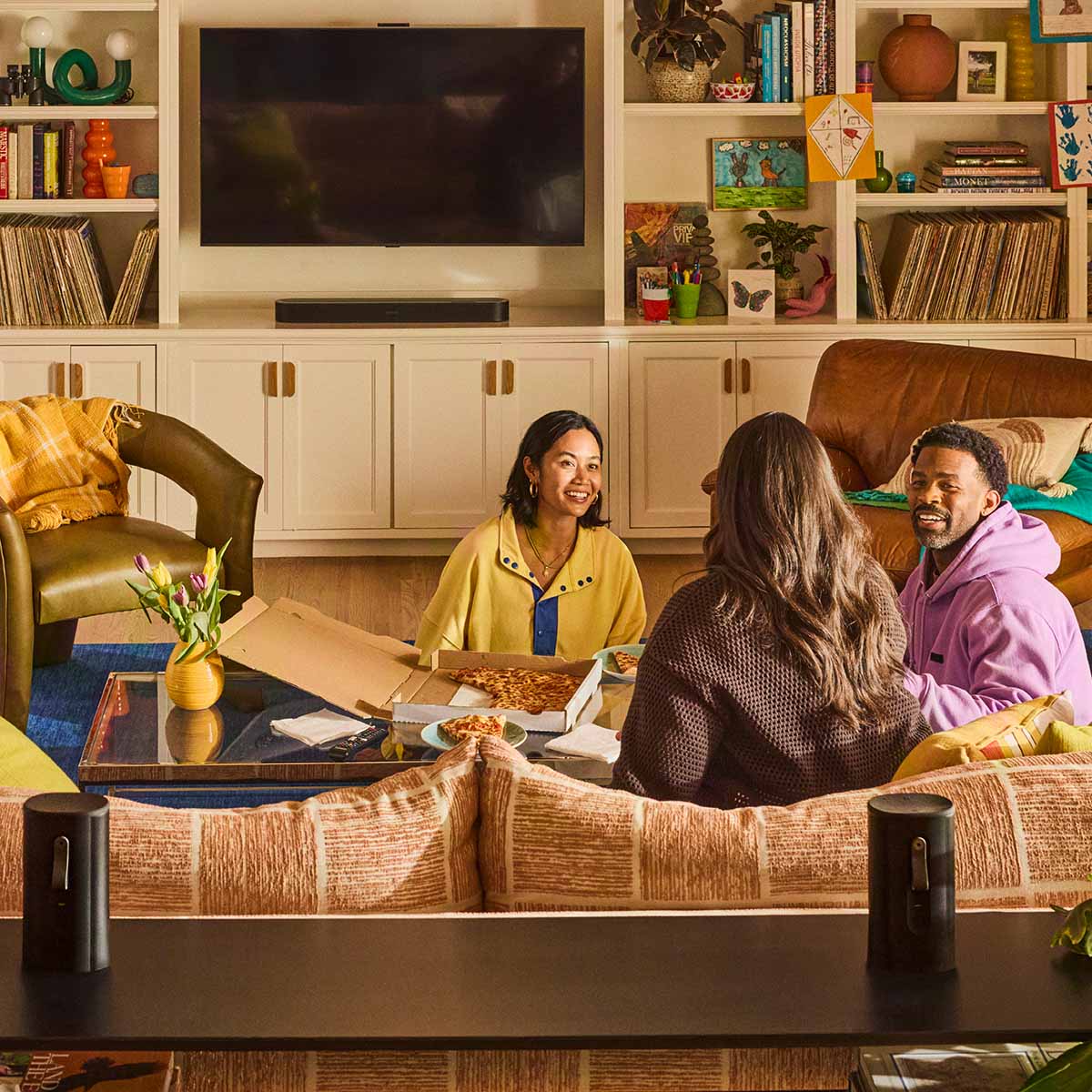 Sonos Beam Ultra Soundbar - lifestyle photo of black soundbar on a cabinet below a TV with group of three eating pizza in foreground