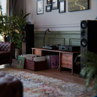 A styled home interior scene showing a wooden record console with a turntable and a Hegel H200 integrated amplifier, flanked by two tall black speakers, with vinyl records in a crate and on shelves. A leather Chesterfield chair, plants, framed wall art, and a patterned rug are visible in a real room setting.