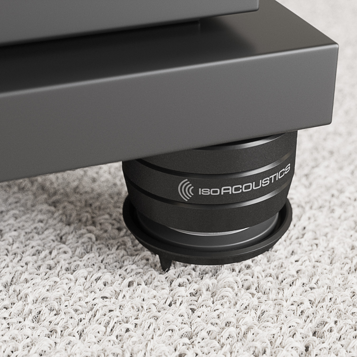 A close-up of a black IsoAcoustics isolation pad supporting a dark gray speaker or equipment base on a white shag carpet.