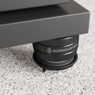 A close-up of a black IsoAcoustics isolation pad supporting a dark gray speaker or equipment base on a white shag carpet.