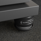 Close-up of a dark gray IsoAcoustics speaker support pad or decoupling base under a speaker cabinet, resting on a textured dark gray carpet.