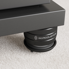 Close-up of a black IsoAcoustics speaker support pad or decoupling pad under a dark gray speaker cabinet, resting on a textured light gray carpet.