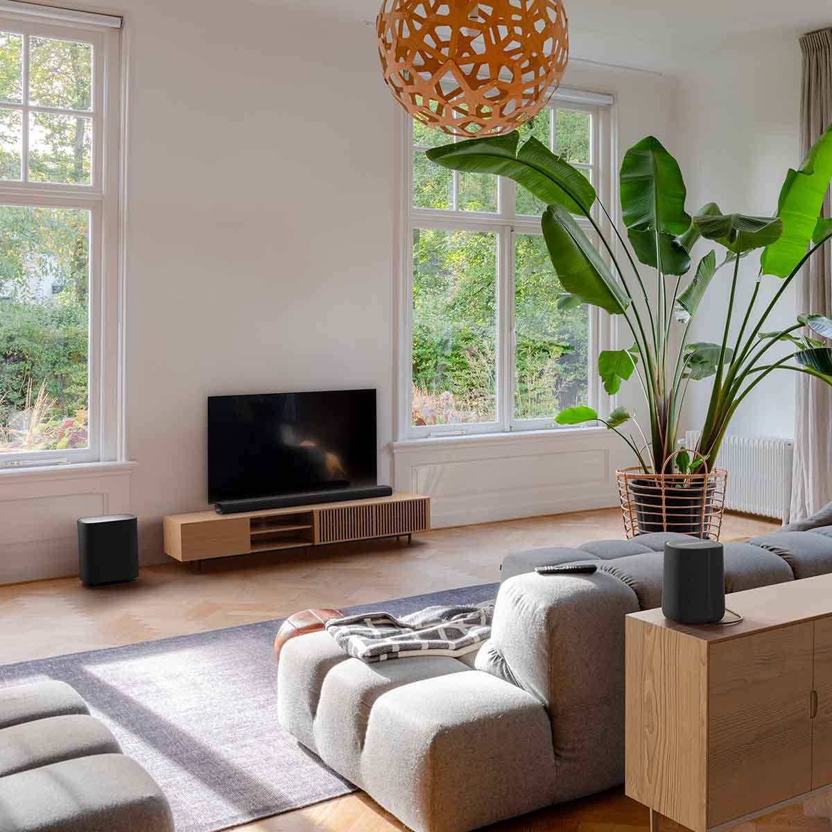 Harman Kardon Enchant 1100 Soudbar lifestyle photo with soundbar in front of TV, sub on floor to left and speaker behind couch