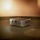A silver metallic Marantz Cinema 50 Series 2 AV Receiver with two rotary knobs and a central circular display, resting on a reflective dark surface against a warm brown gradient background.