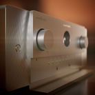 A close-up, angled view of a modern audio receiver or amplifier with a brushed silver metal front panel and large control knobs, set against a warm, blurred brown background.