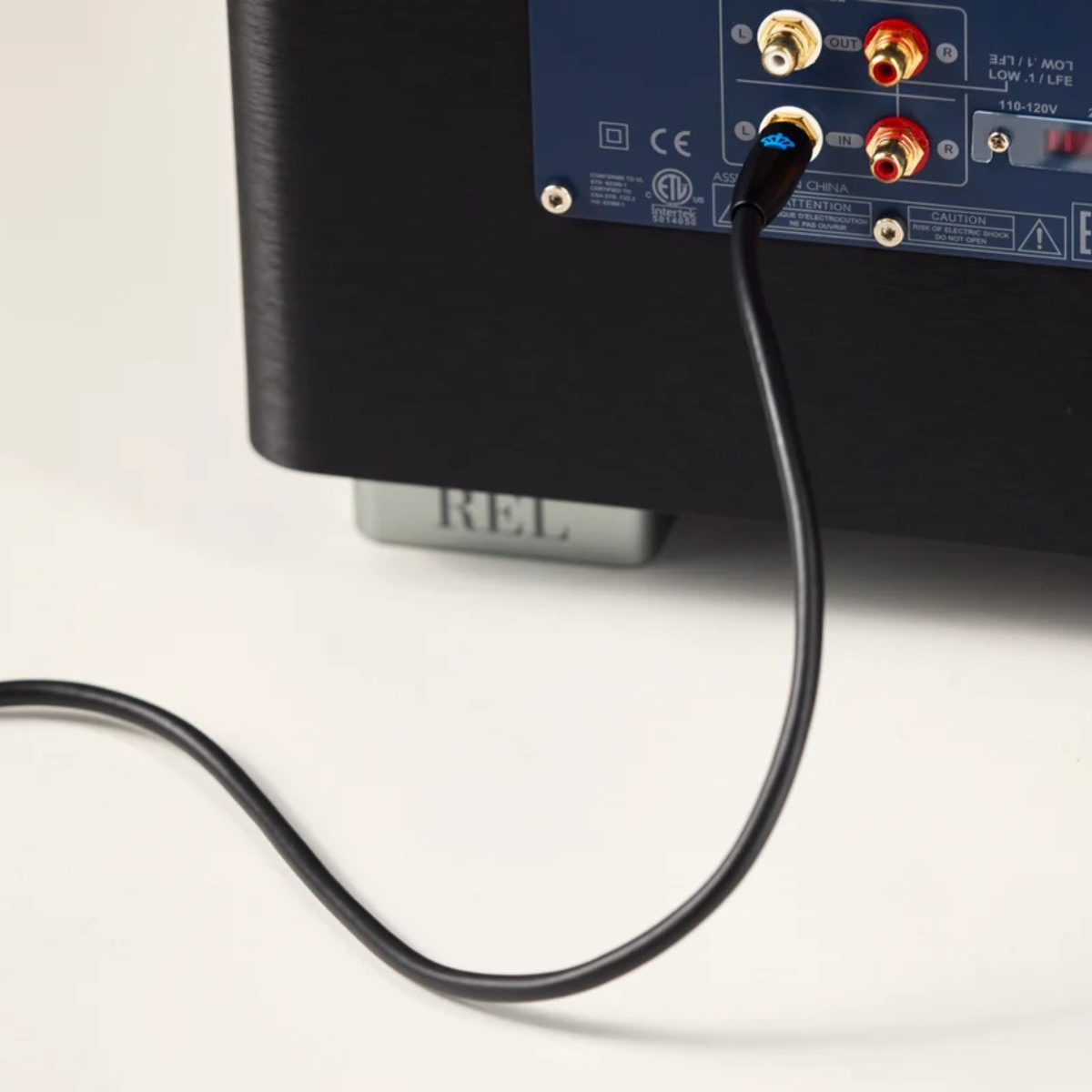 Close-up of a black audio device rear panel with red, white, and gold jacks, with a black REL Acoustics Commander .1/LFE Subwoofer Cable plugged in, on a white background.