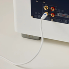 A close-up of a REL Acoustics Commander .1/LFE Subwoofer Cable plugged into the rear panel of a white and black electronic device, with the cable curving downward on a light beige surface.