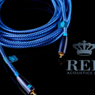 A coiled blue braided REL Acoustics Commodore .1/LFE Subwoofer Cable with gold-plated connectors and small blue brand tags, photographed on a black reflective surface.