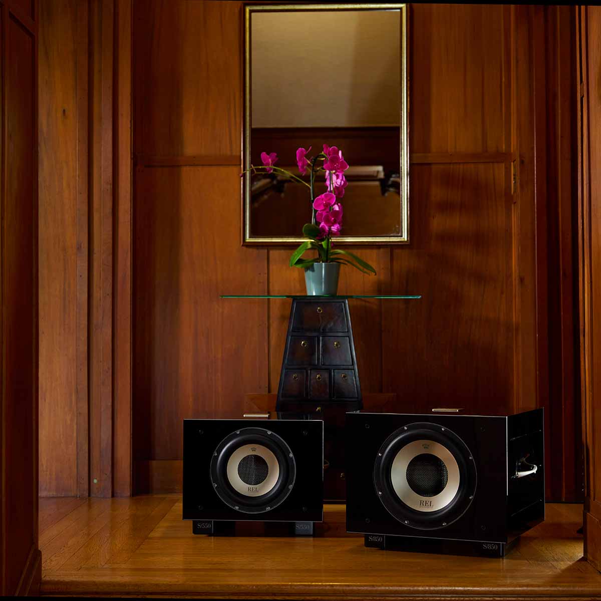 REL Acoustics S/850 Subwoofer - black - lifestyle photo of two subwoofers in front of table with flower in wood panel room no grilles