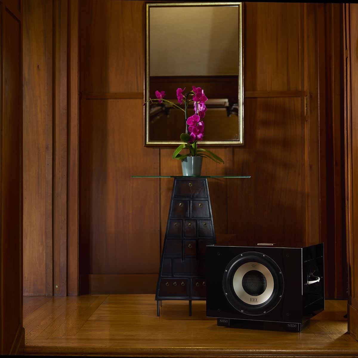 REL Acoustics S/850 Subwoofer - black - lifestyle photo of subwoofer in front of table with flower in wood panel room no grille