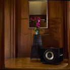 REL Acoustics S/850 Subwoofer - black - lifestyle photo of subwoofer in front of table with flower in wood panel room no grille