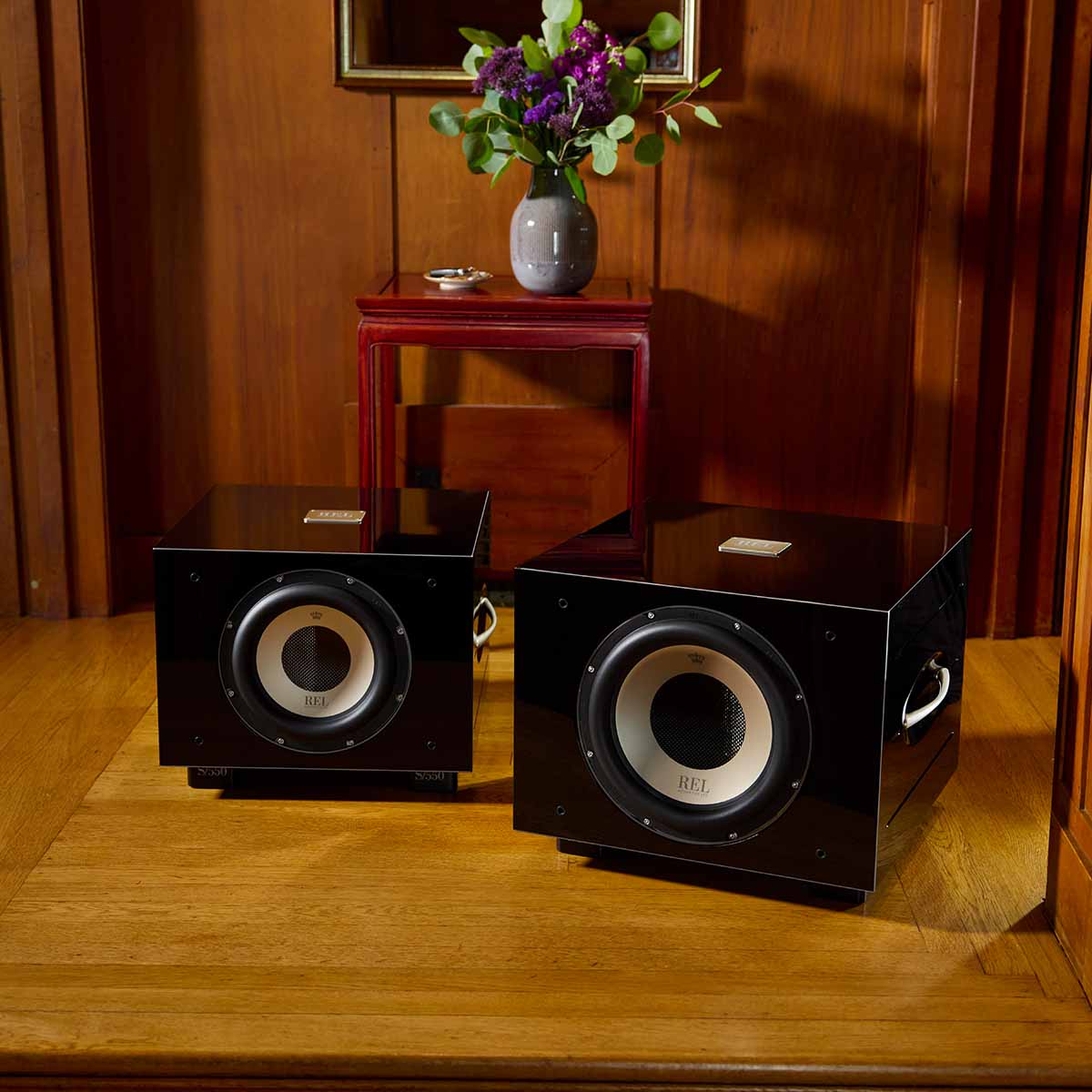 REL Acoustics S/850 Subwoofer - black - lifestyle photo of two subwoofers in front of table with flower in wood panel room no grilles