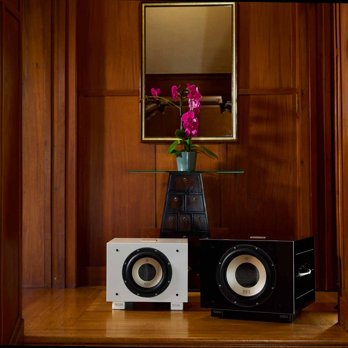 REL Acoustics S/850 Subwoofer - black and white - lifestyle photo of two subwoofers in front of table with flower in wood panel room no grilles