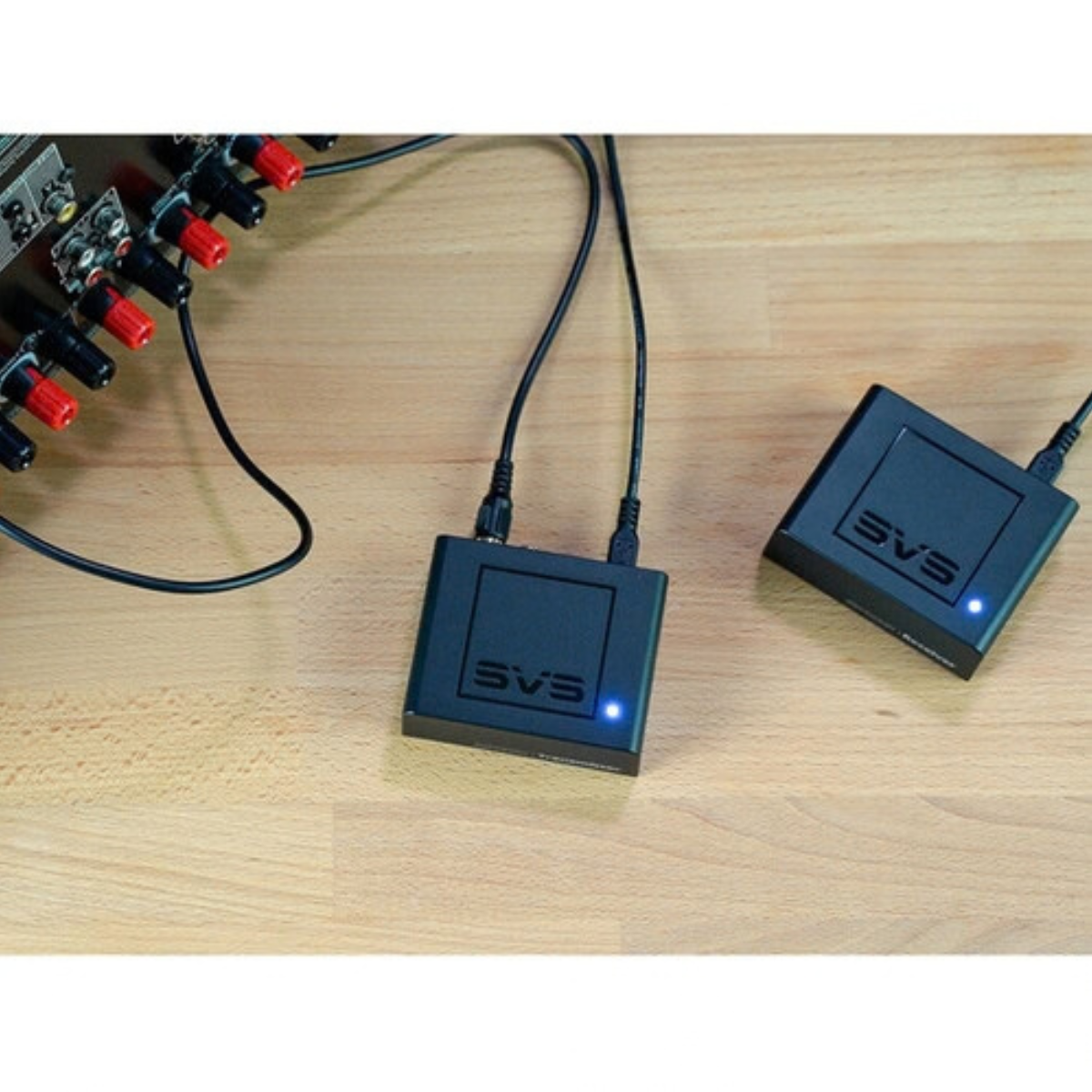 Two black SVS audio power adapters with blue LED indicators, connected to cables and placed on a light wood surface next to an audio device.