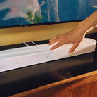 Sonos Beam Ultra Soundbar - lifestyle photo closeup of white soundbar on a cabinet in a living room with hand pressing a button on the bar