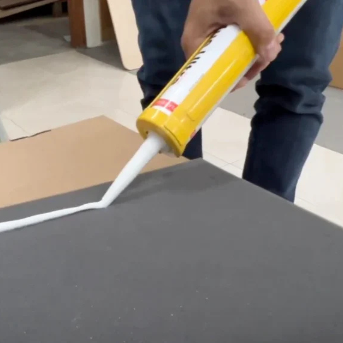 A person using a yellow and white caulking gun to apply a thick Vicoustic Flexi Glue Ultra High Tack Glue onto a dark gray surface.