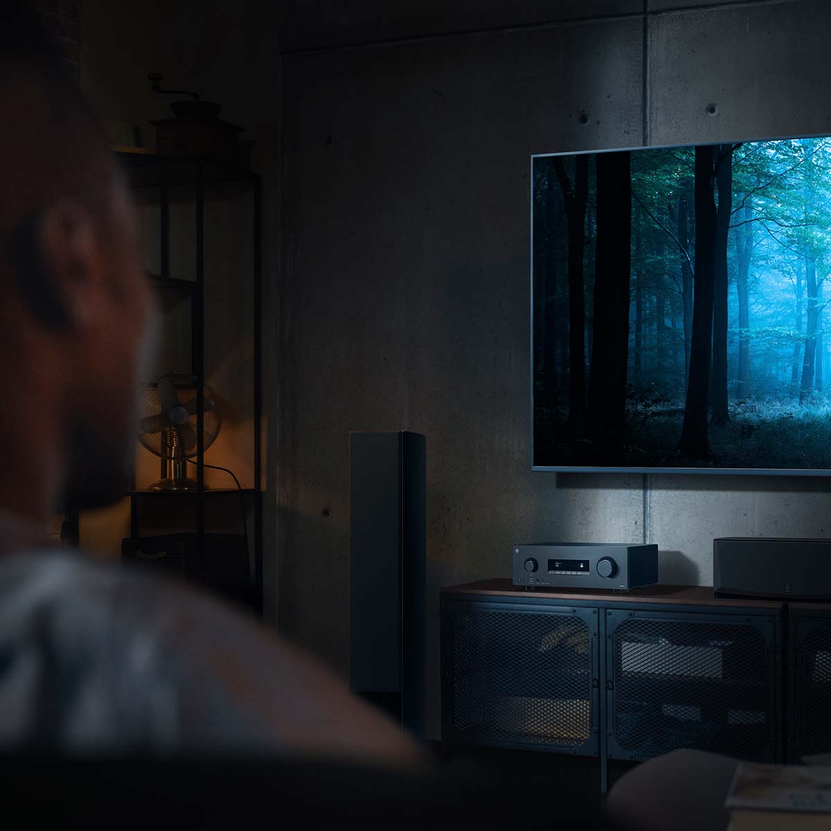 Yamaha RX300A 5.2-Channel AV Receiver - lifestyle image with dark lighting of unit on cabinet front view angled left below TV with out of focus man in foreground