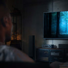 Yamaha RX300A 5.2-Channel AV Receiver - lifestyle image with dark lighting of unit on cabinet front view angled left below TV with out of focus man in foreground
